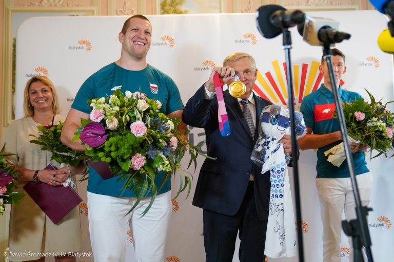 Na białej ściance, z logiem wschodzącego Białegostoku, po lewej stronie stoi młociarz Wojciech Nowicki, trzyma w rękachkwiaty, z prawej prezydent Białegostoku, który trzyma złoty medal