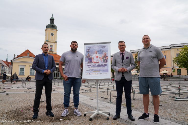 Czterech mężczyzn stoi na Rynku Kościuszki w Białymstoku, między nimi tablica z plakatem informującym o imprezie lekkoatletycznej. W tle widać ratusz