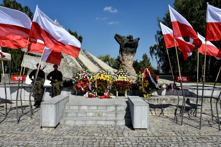 Dwaj żołnierze stoją przy pomniku obrońców miasta, przed pomnikiem stoją wieńce kwiatów, na wietrze trzepoczą biało-czerwone flagi