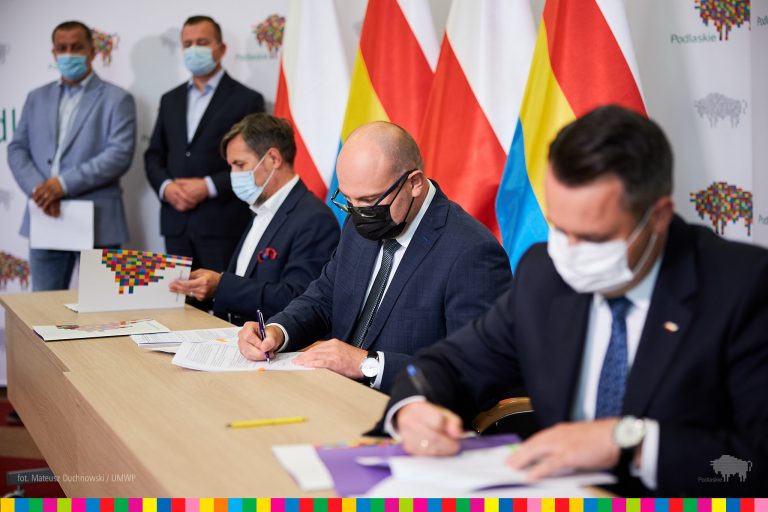 Trzech mężczyzn siedzi przy biurku i podpisuje umowy . W tel stoi jeszcze dwóch mężczyzn oraz flagi Polski i województwa podlaskiego