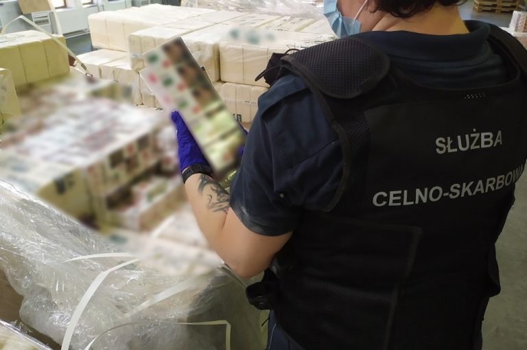 Funkcjonariuszka Służby Celno-Skarbowej trzyma karton nielegalnych papierosów. W tle większa ilość papierosów na paletach.