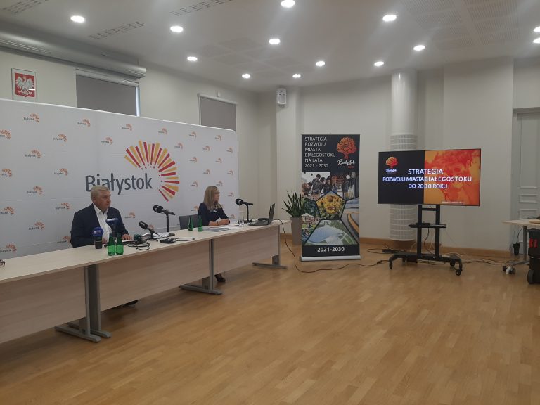 Sala konferencyjna, z lewej strony stoi ścianka z napisem Bialystok, na jej tle przy stole siedzi prezydent Białegostoku oraz kobieta, na wprost stoi stand, lekko na prawo telewizor z wyświetlonym napisem: Strategia Białegostoku