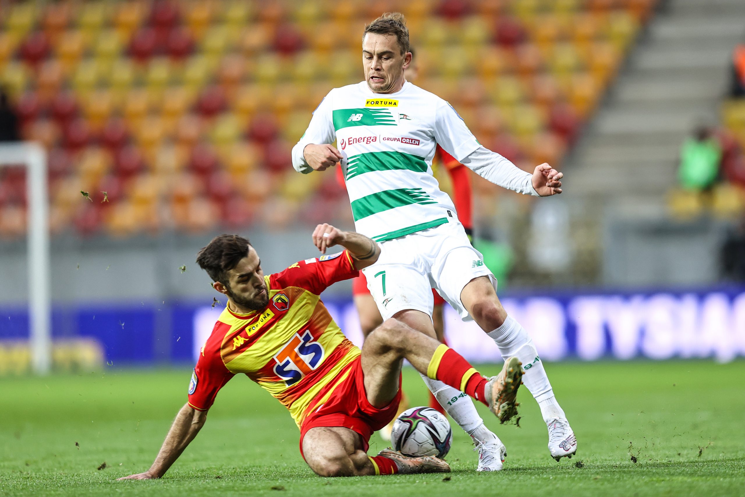 Na murawie siedzi piłkarz w żółto - czerwonym stroju i próbuje wybić piłkę zawodnikowi w biało - zielonym stroju, który stoi nad nim