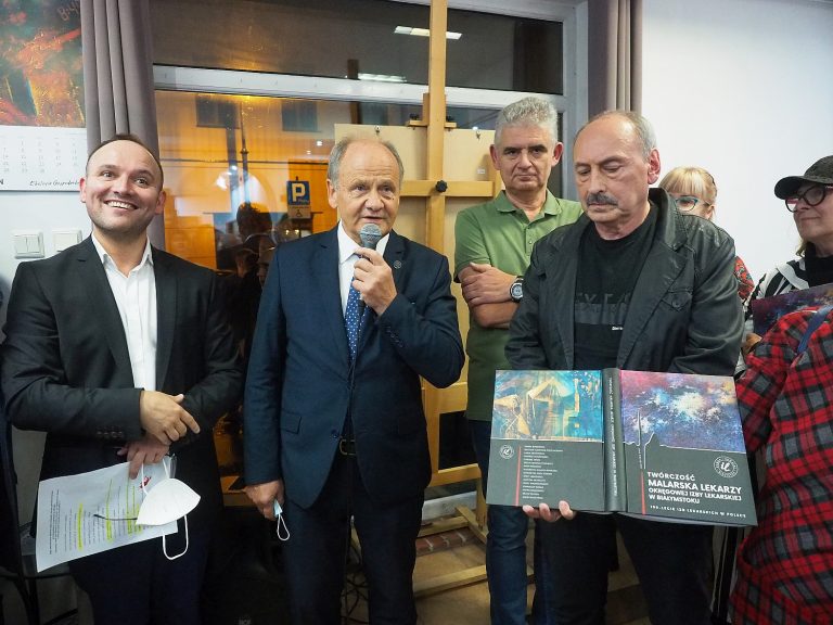 Wernisaż wystawy. Grupka osób stroi obok siebie. Jeden z mężczyznmówi do mikrofonu, drugi prezentuje album fotograficzny