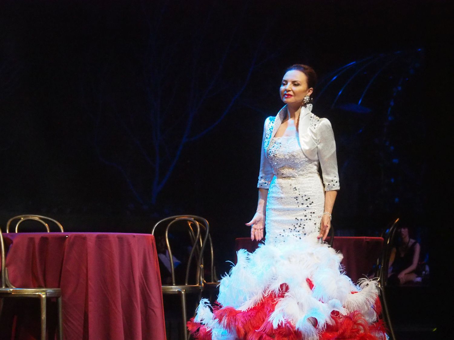 Kobieta w białej sukni stoi na scenie. Obok niej stoi okrągły stolik nakryty czerwonym materiałem