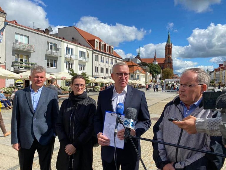 Cztery osoby stoją na Rynku Kościuszki w Białymstoku. Za nimi widać kamienicę oraz katedrę białostocką. Mężczyzna stojący po środku mówi do mikrofonu