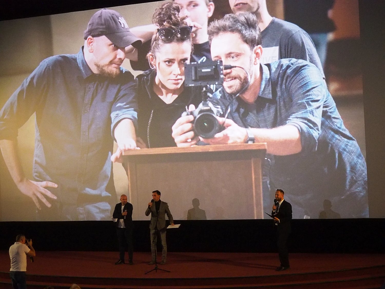 Na scenie stoi trzech mężczyzn, za nimi na ekranie kinowym wyświetlany jest kadr z filmu