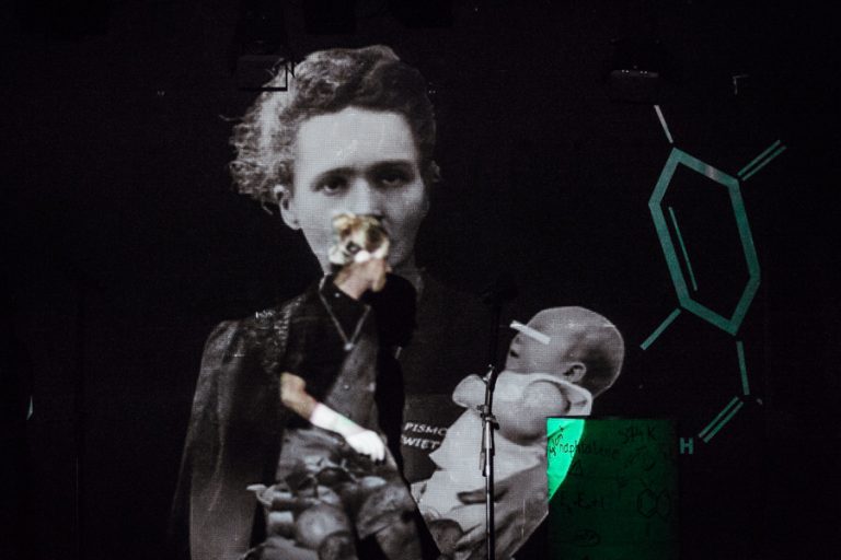 Kobieta na scenie, w tle wizualizacja przedstawiająca Marię Skłodowską-Curie