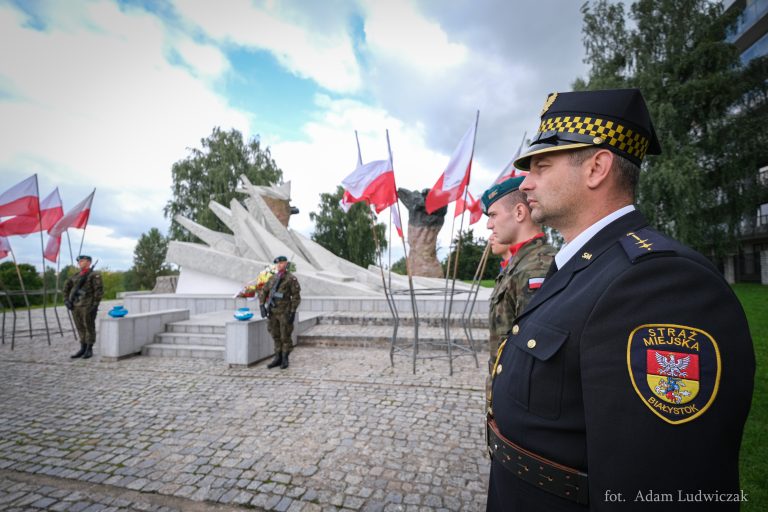 żołnierze i strażnicy miejscy stoją przy pomniku obrońców miasta, przed pomnikiem stoją znicze, na wietrze trzepoczą biało-czerwone flagi