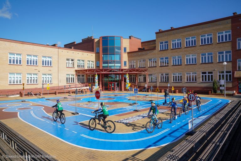 Plac przed budynkiem szkoły, na którym urządzono miasteczko ruchu drogowego. Są niebieskie jezdnie, stoją znaki drogowe, na rowerach jeździ kilkoro uczniów