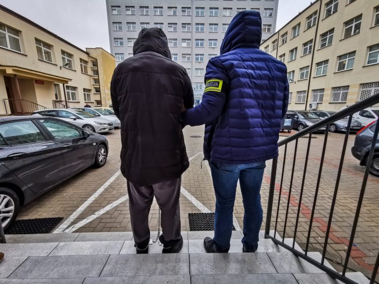 Policjant trzyma za ramię skutego kajdankami mężczyznę