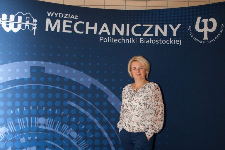 Kobieta w jasnej bluzce stoi przed ścianką z napisem Wydział Mechaniczny PB