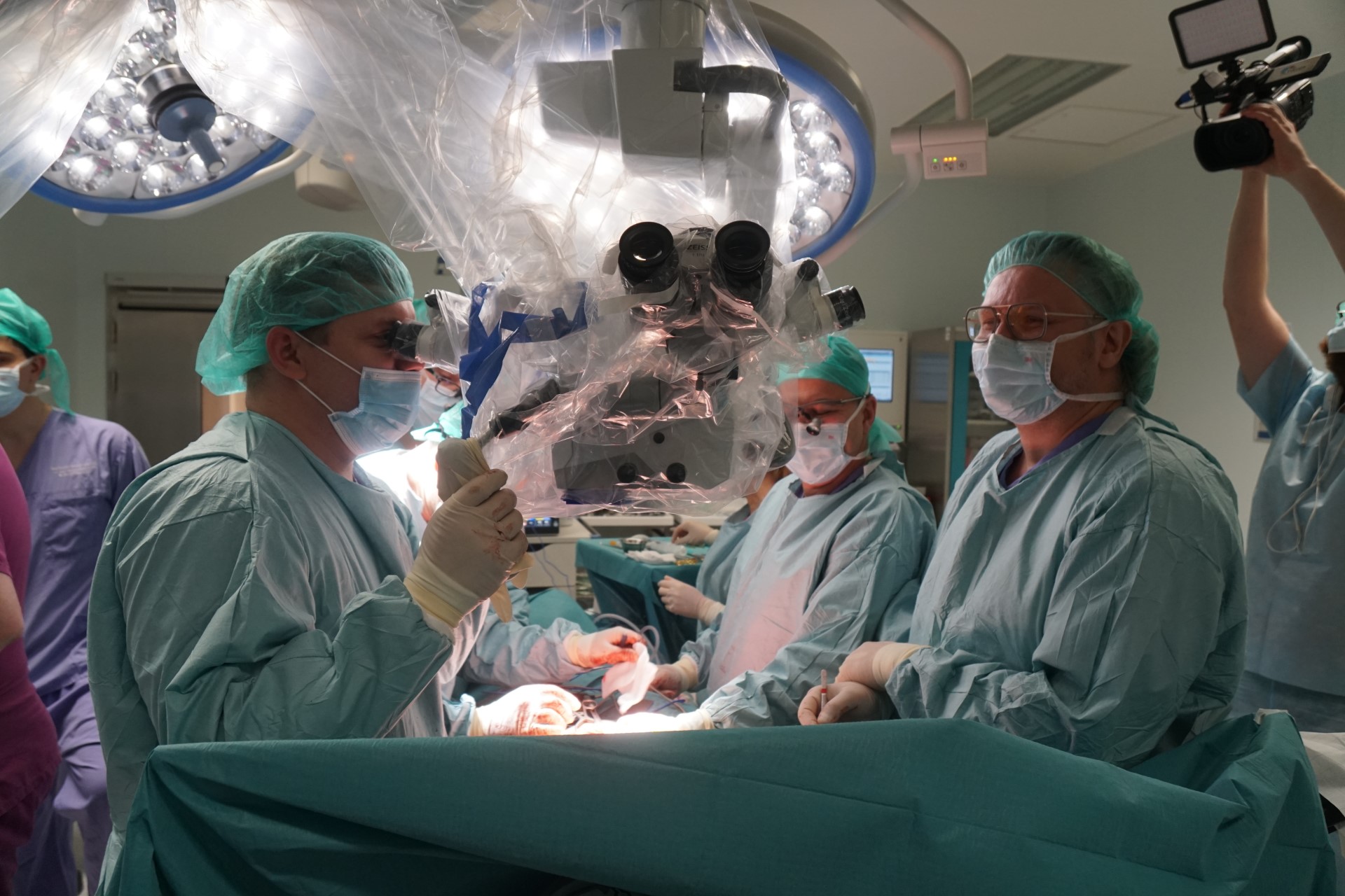 Trwa operacja, przy stole w zielonych fartuchach i czepkach na głowie stoją lekarze. Nad pacjentką, na wysokości jej klatki piersiowej wisi mikroskop, przez który patrzy lekarz znajdujący się po lewej stronie fotografii