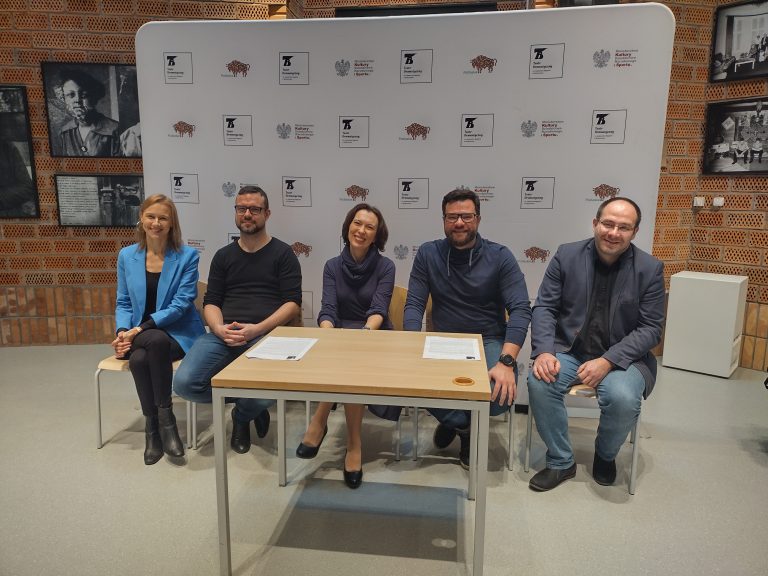 NA tle ścianki Teatru Dramatycznego siedzi pięć osób, przed nimi stoi stolik