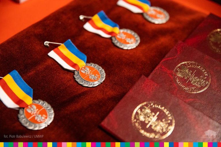 Na czerwonym welurze leżą medale, z biało-czerwono-żółto-niebieskim paskiem i metalową, czerwoną rozetką, na której wytłoczone jest godło województwa podlaskiego