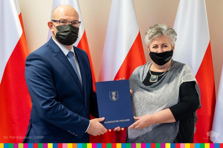 Mężczyzna w ciemny garniturze, w okularach i maseczce na twarzy przekazuje nominację dyrektorską kobiecie w czarno -szarym ubraniu , która ma maseczkę na twarzy