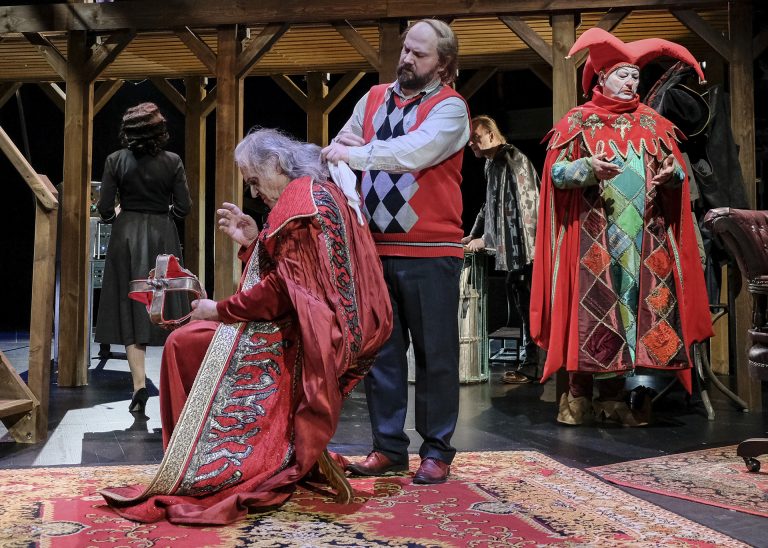 Scena teatralna, na podłodze leży czerwony, w roślinne wzory dywan, z lewej strony na krześle siedzi mężczyzna w czerwonym królewskim stroju, ma na sobie płaszcz heraldyczny, jego siwe włosy sięgające do szyi czesze mężczyzna, który stoi nad nim. To jest garderobiany, który ma na bok zaczesane włosy i jest ubrany w czerwony sweter w czarne romby na brzuchu. Z prawej strony stoi mężczyzna przebrany za błazna, ma kolorową pelerynę, w dominującym czerwonym kolorze, z przodu są zielone i bordowe romby. Ma na biało umalowaną twarz