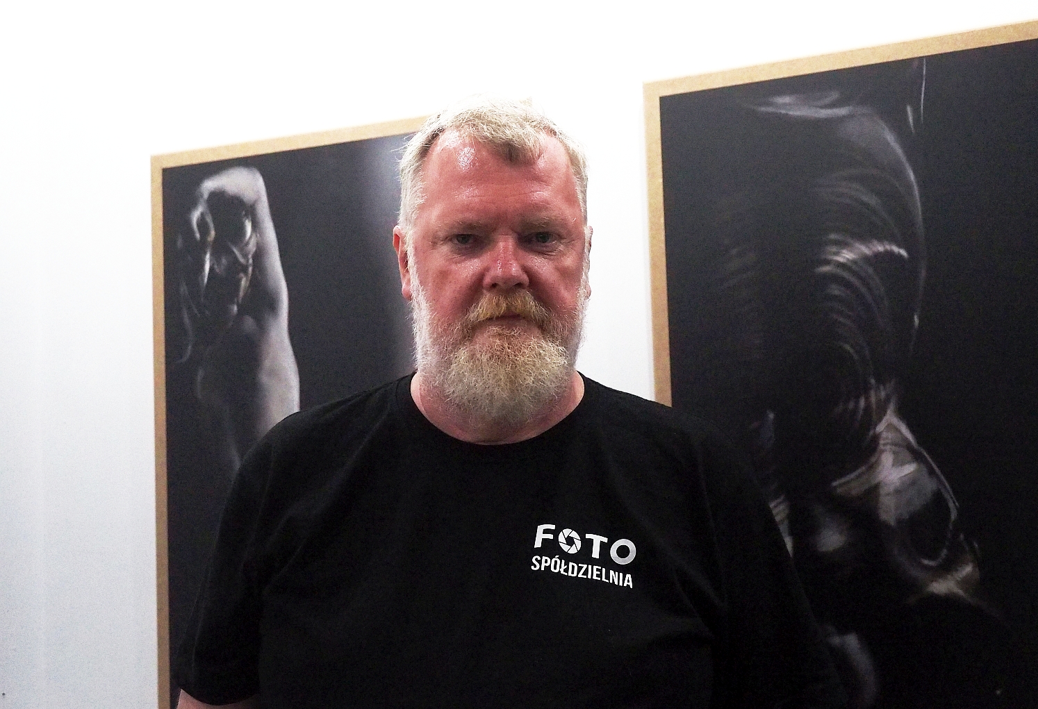 Mężczyzna w czarnym tiszercie, z siwą brodą stoi na tle ciemnych zdjęć, które powieszone są na białej ścianie