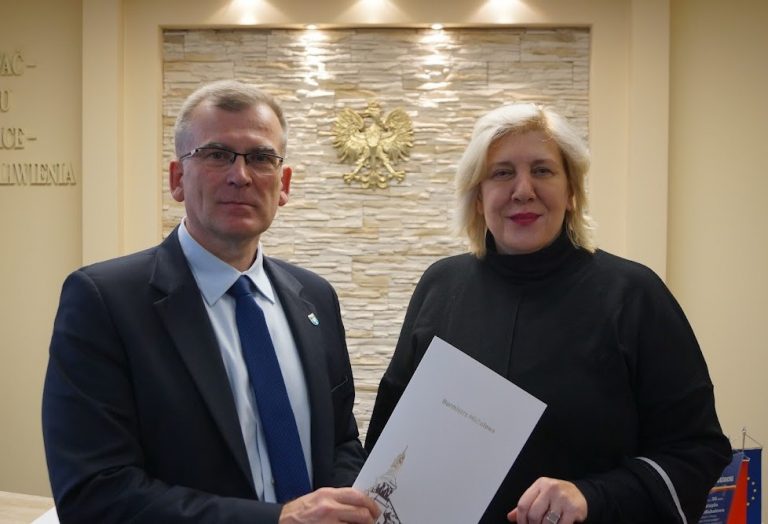 Na tle kremowej ściany, na której wisi złote godło Polski stoi mężczyzna w ciemnym garniturze i kobieta w czarnej bluzie. Mężczyzna trzyma przed sobą w ręku białą teczkę
