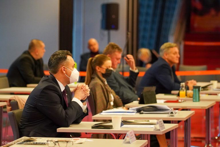 Przy pojedynczych stolikach siedzą radni. NA pierwszym planie siedzi mężczyzna w ciemnym garniturze, obok niego kobieta