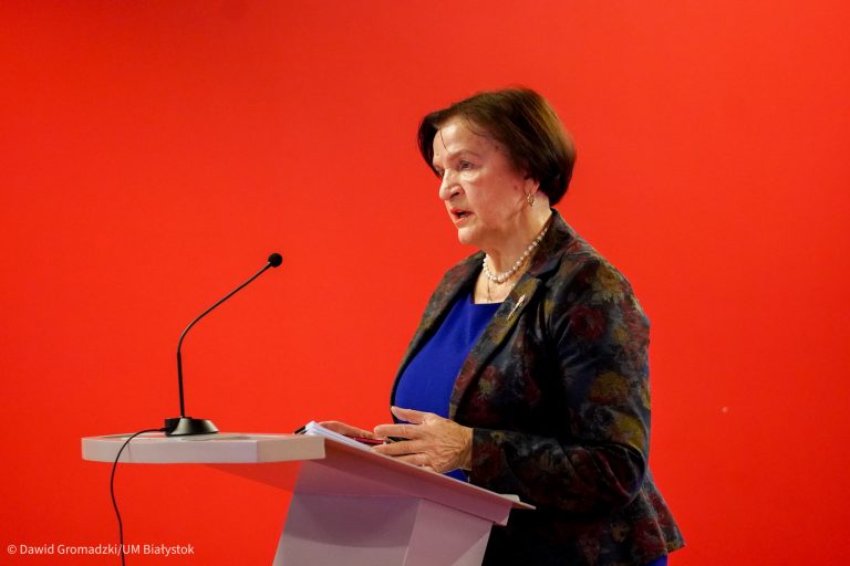 Na tle czerwonej ścianie, przy mównicy stoi kobieta, która przemawia. Jest w średnim wieku, ma krótkie ciemne włosy i ciemny wzorzysty żakiet, pod którym ma w kolorze kobaltowym bluzkę