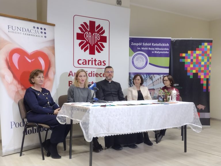 Przy stole nakrytym białym obrusem siedzi pięć osób. Po dwie kobiety po bokach, w środku ksiądz. Za nimi stoją banery; Fundacji Rodziny Czarneckich, Caritas Polska, województwa podlaskiego