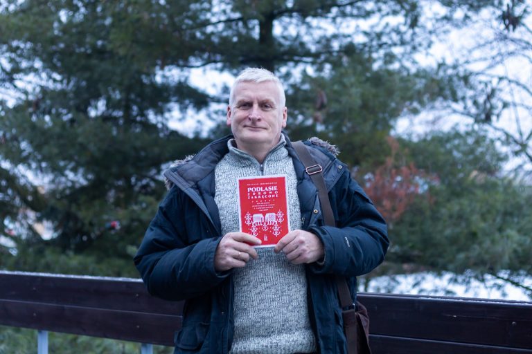 Siwy mężczyzna stoi przy barierce, w rękach ma książkę