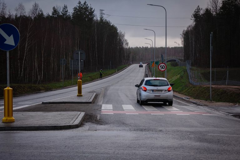 Droga poza terenem zabudowanym, po której jedzie jasny samochód. Auto widoczne jest od tyłu