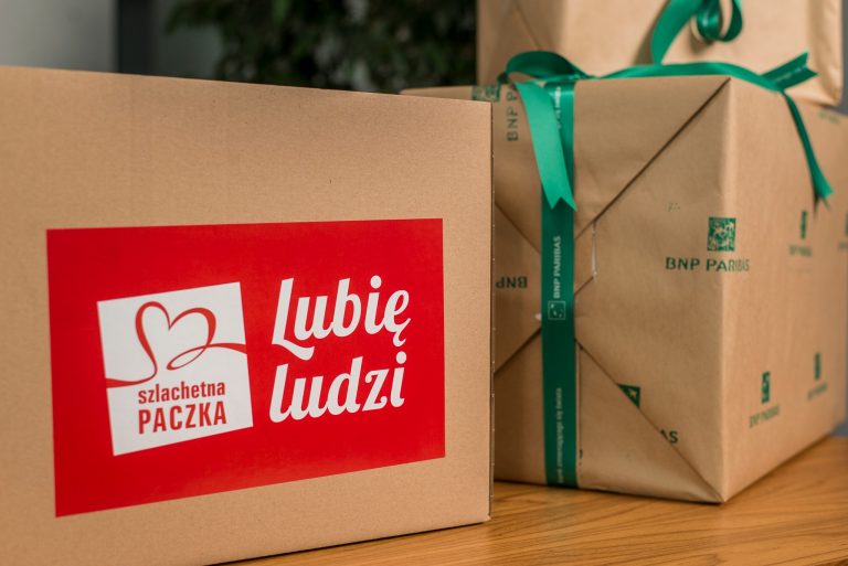Dwa prezenty, w prostokątnych pudełkach, zawinięte w brązowy papier. Na pudełku z lewej strony jest przyklejona czerwona naklejka z białym napisem: Lubię ludzi, na prezencie z prawej zawiązana jest zielona wstążka