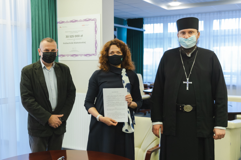 W pokoju na tle jasnej ścianki działkowej stoją trzy osoby; od lewej mężczyzna, po środku kobieta, która trzyma przed sobą podpisaną umowę.z lewej duchowny