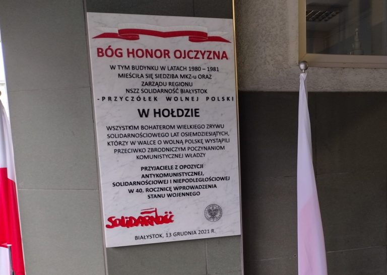 Biała, kamienna tablica z czarnymi napisami wisi na zewnętrznej ścianie. Po obu stronach tablicy znajdują się biało-czerwone flagi