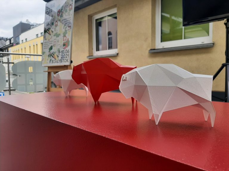Na czerwonym stole stoją trzy plastikowe żubry; biały, czerwony, biały. W oddali, z lewej strony na sztaludze stoi plakat, z prawej telewizor