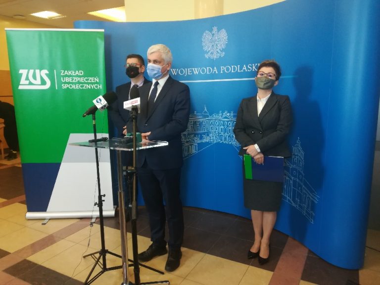 Trzy osoby, kobieta i dwóch mężczyzn stoi na tle banera Podlaskiego Urzędu Wojewódzkiego podczas konferencji prasowej. To: Bohdan Paszkowski – wojewoda podlaski, Piotr Jurczuk – zastępca dyrektora Izby Administracji Skarbowej w Białymstoku, Beata Pawlińska-Wiak – dyrektor oddziału Zakładu Ubezpieczeń Społecznych w Warszawie