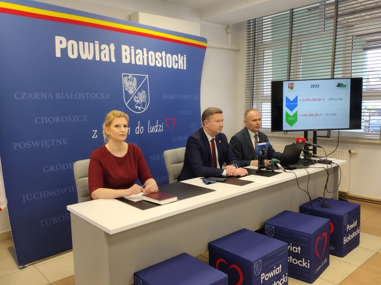 Przy jasnym stole siedzą trzy osoby na tle niebieskiej ścianki z białym napisem: Powiat Białostocki. Od lewej strony siedzi kobieta w bordowej sukience, po środku i z lewej strony mężczyźni w ciemnych garniturach. Z prawej strony stoi monitor, na którym wyświetlana prezentacja