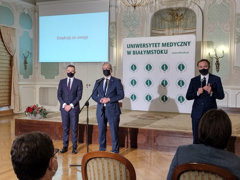 W auli przed podwyższeniem , z lewej strony stoi dwóch mężczyzn. Za nimi znajduje się rzutnik, na którym na białym tle jest wyświetlony czerwony napis: Dziękuję za uwagę. Od mężczyzn z lewej strony w pewny odstępie znajduje się kolejny mężczyzna, stoi on bardziej z prawej strony i mówi do mikrofonu. W tle widać białą ściankę z zielonym napisem: Uniwersytet Medyczny w Białymstoku