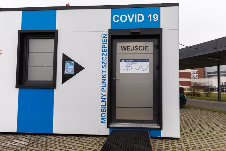 Wejście do konternerowego mobilnego punktu szczepień przeciwko COVID-19. Fragment punktu jest w biało-niebieskie pionowe pasy. Drzwi wejściowe są szare, nad nimi znajduje się biały napis na niebieskim tle: COVID-19