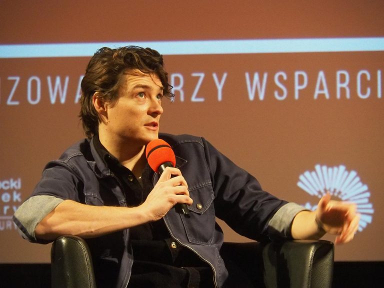 Młody mężczyzna w jeansowej koszulki siedzi w fotelu podczas spotkania i mówi do mikrofonu. Za nim fragment pomarańczowego tła, które wyświetlane jest na kinowym ekranie