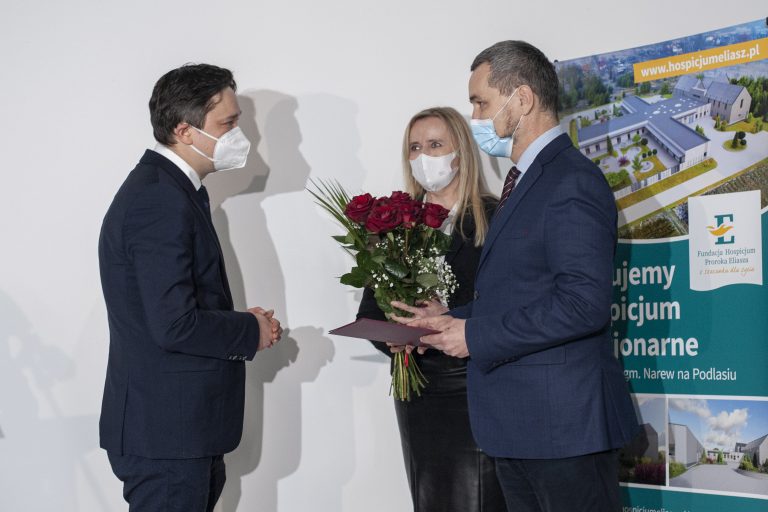 Z lewej strony, bokiem stoi mężczyzna w białej maseczce ochronnej zasłaniające noc i usta, ubrany jest w ciemno-granatowy garnitur, ma ciemne włosy. Z prawej strony w głębi zdjęcia znajduje się kobieta, która ma blond włosy, również maseczkę ochronną, jest w ciemnym stroju i trzyma przed sobą bukiet czerwonych róż. Obok niej stoi mężczyzna w maseczce ochronnej i ciemno-granatowym garniturze