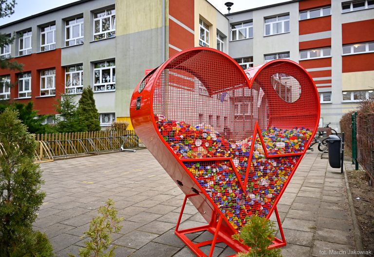Przed budynkiem szkoły stoi metalowy pojemnik w kształcie serca na plastikowe nakrętki