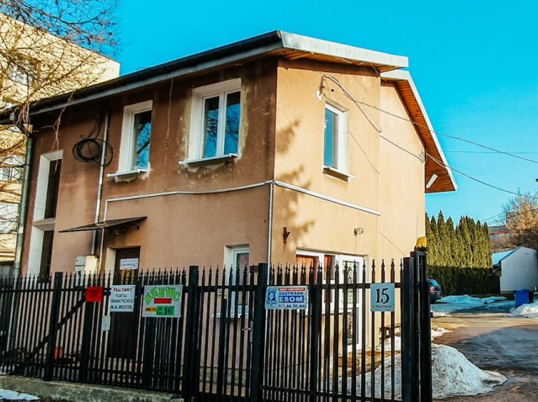 Jest słoneczny, bezchmurny dzień. Na zdjęciu widoczny jest jasno-pomarańczowy budynek z oknami. Przed budynkiem jest brązowa, żelazna brama