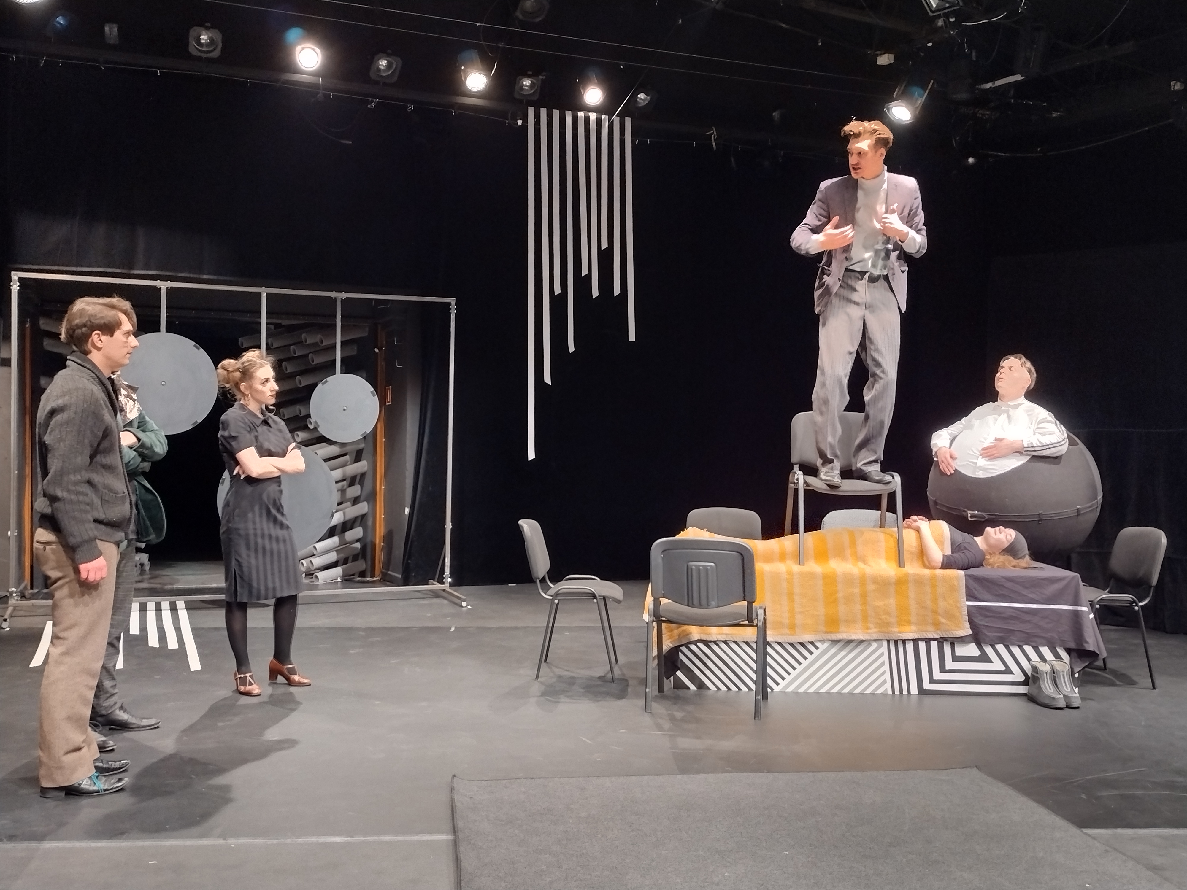 Scena z "Tanga" Sławomira Mrozka". Szaro-czarna scenografia, z lewej strony stoją trzy osoby, dwóch aktorów i aktorka, która ma założone ręce i patrzy na mężczyznę, taj jak pozostali, który stoi na stole