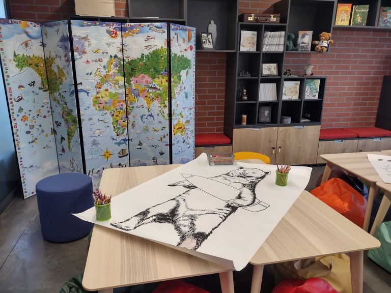 Sala edukacyjna, na stole leży dużego formatu kartka, na której jest niedźwiedź do pokolorowania. W oddali stoi parawan przegrodowy, który jest mapą. Obok parawanu są półki zawieszona na ceglasto-czerwonej ścianie