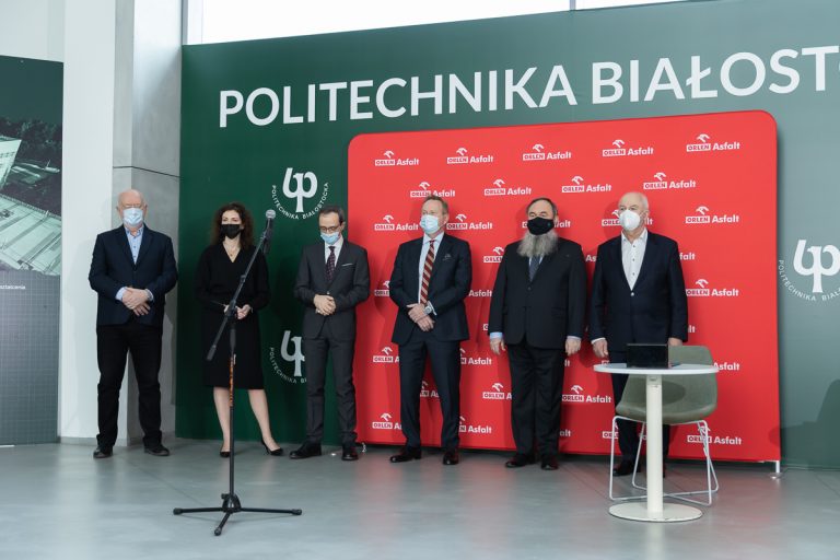 Na tle zielonej ścianki z białym napisem: Politechnika Białostocka, ustawiono mniejszą czerwoną ściankę z białymi napisami Orlen Asfalt, na ich tle stoi sześć osób. Przed nimi jest mikrofon na statywie oraz z prawej strony, biały, okrągły stolik