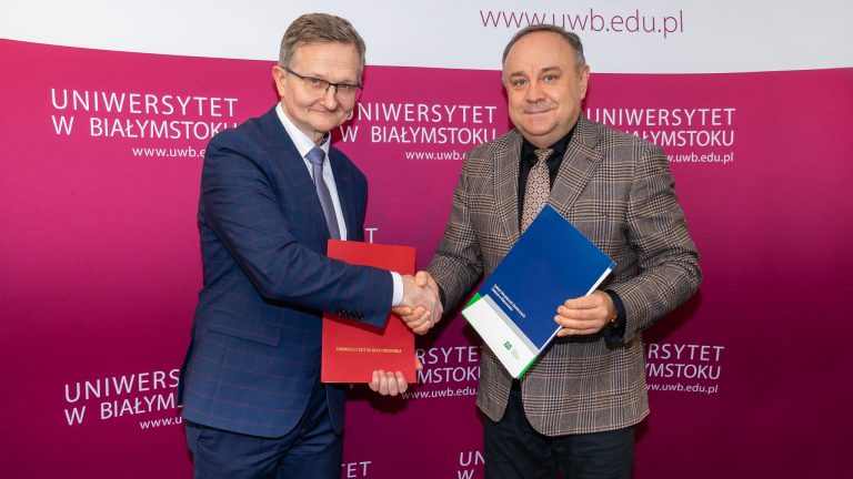 Na różowej ściance z białym napisem: Uniwersytet w Białymstoku stoi dwóch mężczyzn, którzy wzajemnie ściskają sobie dłonie