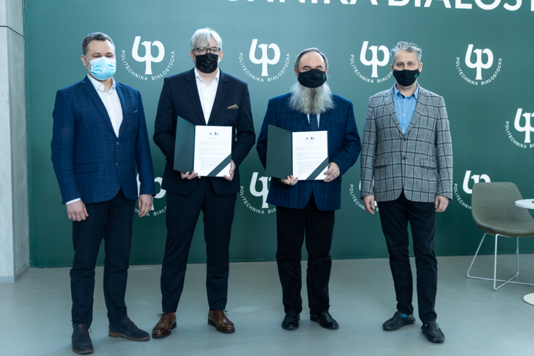 Na tle zielonej ścianki w znaki Politechniki Białostockiej stoi czterech mężczyzn, dwóch stojących w środku trzyma przed sobą podpisane umowy