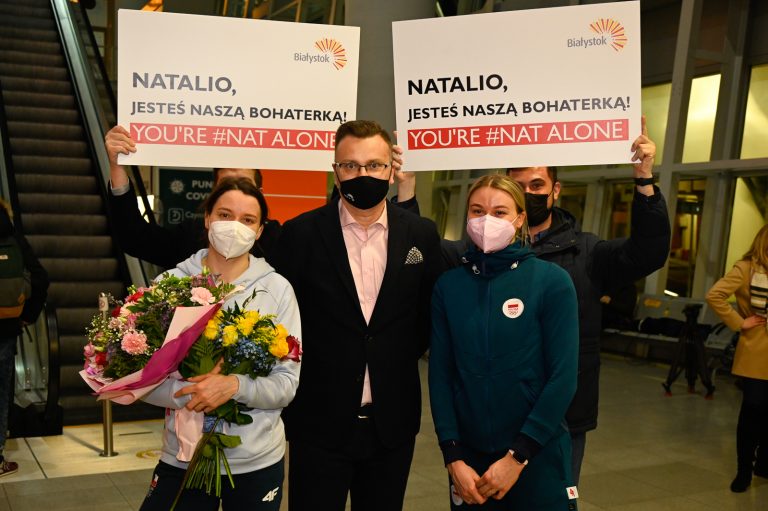 Pomiędzy dwiema kobietami w sportowych strojach stoi mężczyzna w ciemnej marynarce i jasno różowej koszuli . Za nimi stoją dwie osoby, które trzymają tabliczki z napisami: Natalio, jesteś naszą bohaterką. Pod spodem na czerwonym pasku biały napis : You're #Nat Alone