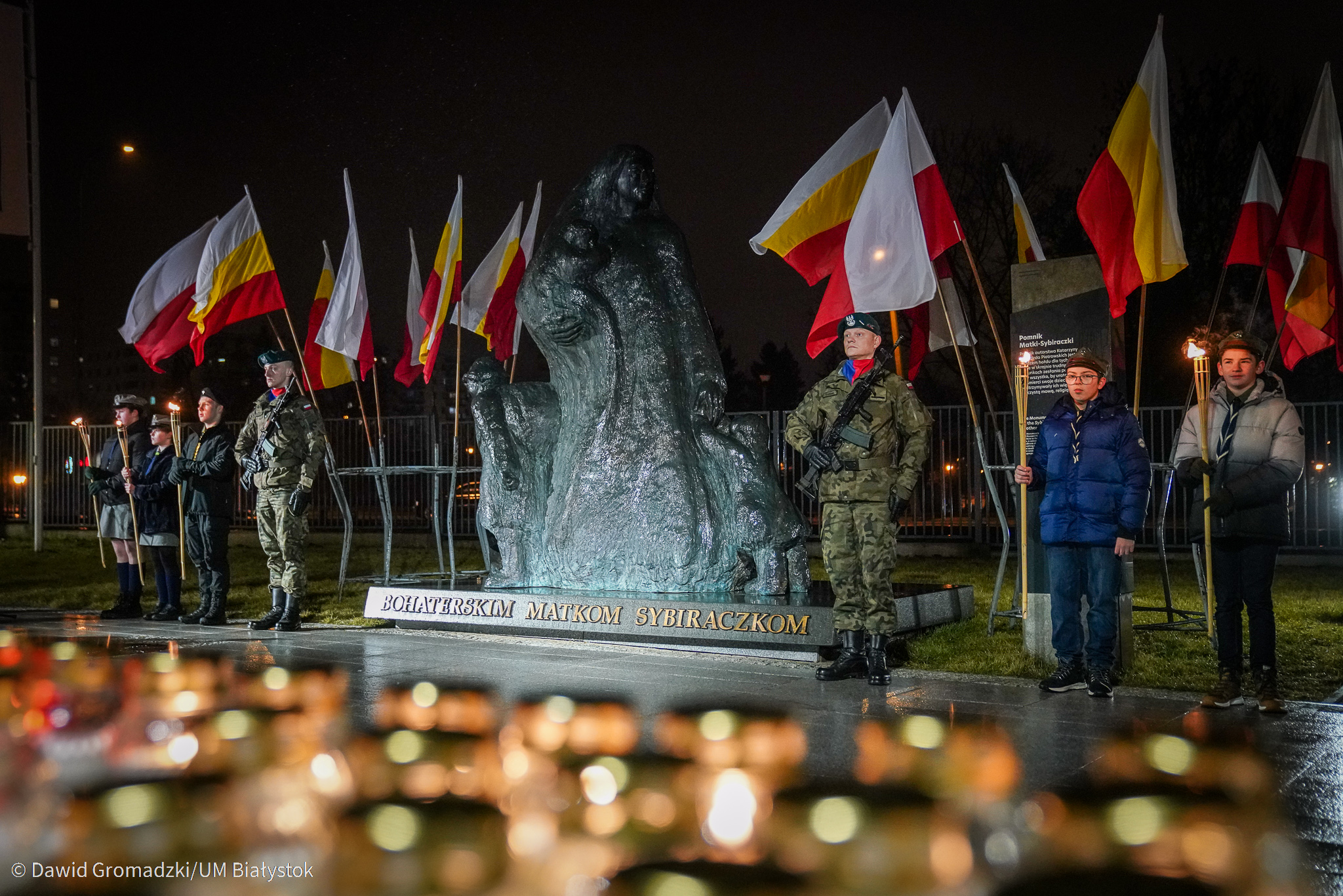 Na pierwszym planie palą się znicze. W oddali stoi pomnik Bohaterskich Matek Sybiraczek. Po obu stronach monumentu znajdują się harcerze, którzy stoją na baczność. Nad ich głowami powiewają flagi Polski oraz Białegostoku, które umieszczone są w metalowych stojakach