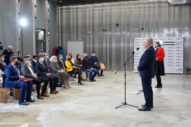 W sali, która w ogólnym odbiorze jest metalowo-szara, po lewej stronie na krzesłach siedzą różne osoby; dorośli i młodzież, z prawej przy mikrofonie stoi mężczyzna, obok niego znajduje się kobita w czerwonej marynarce