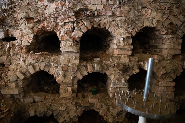 Fragment nisz grzebalnych w supraskich katakumbach. Widać sześć nisz w ścianie z cegły, która jest w złym stanie. Z boku, z prawej strony stoi stolik-świecznik (panichidnik)