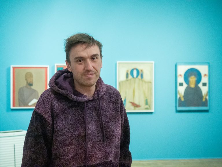 Mężczyzna na tle niebieskiej ściany z zawieszonymi na niej pracami